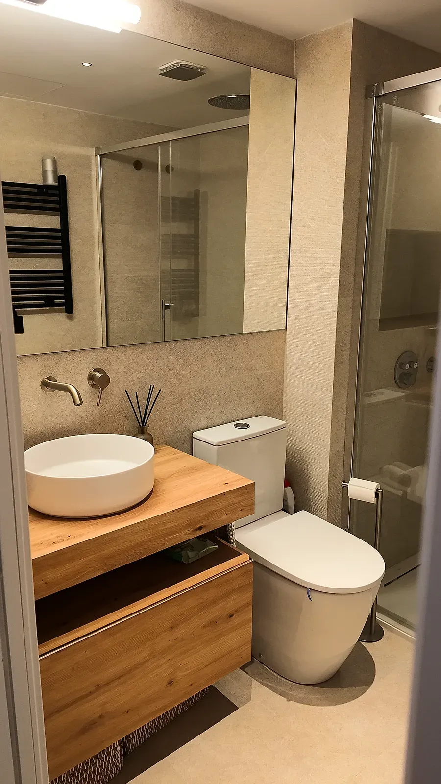 Baño completo en Castelldefels con lavabo redondo, mueble de madera, ducha acristalada y toallero negro